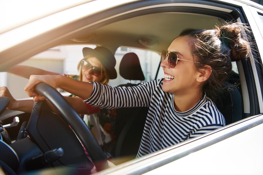 Two attractive young girlfriends wearing sunglasses talking and laughing together while driving in a GoTo Flex Lease car through the city on a sunny day