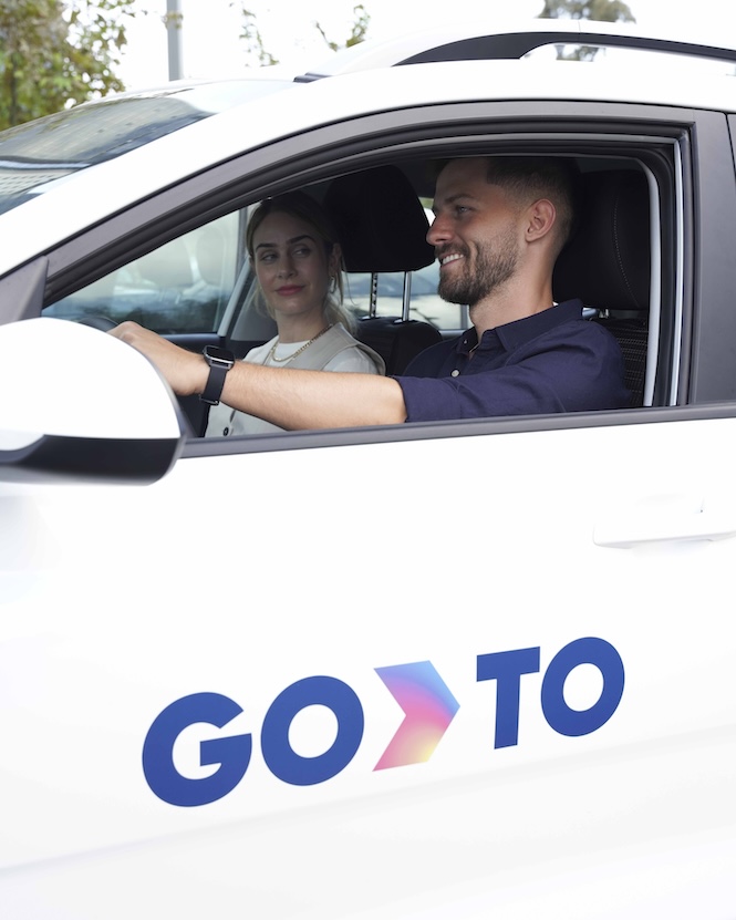 A man and a woman sitting in a GoTo shared vehicle, with the man driving and smiling. The side of the white car displays the GoTo logo in bold blue letters with a gradient arrow.