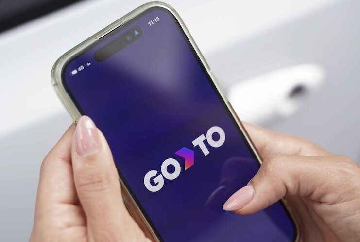 A person holding a smartphone with the GoTo app open on the screen, ready to use. The phone displays the GoTo logo, and the individual stands near a vehicle, hinting at the app's use for managing vehicle.