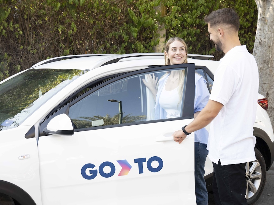 A man opening the door for a woman as she gets into a GoTo shared vehicle. Both are smiling, with the GoTo logo clearly visible on the side of the car. The scene suggests a friendly interaction facilitated by the GoTo car-sharing platform.