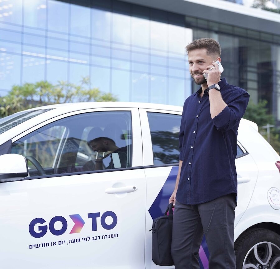 A man stands next to a GoTo shared vehicle, talking on his phone while holding a black briefcase. The white car features the GoTo logo and text indicating vehicle rentals by the hour, day, or month. The background shows a modern office building, emphasizing the business context of GoTo's services for professionals.