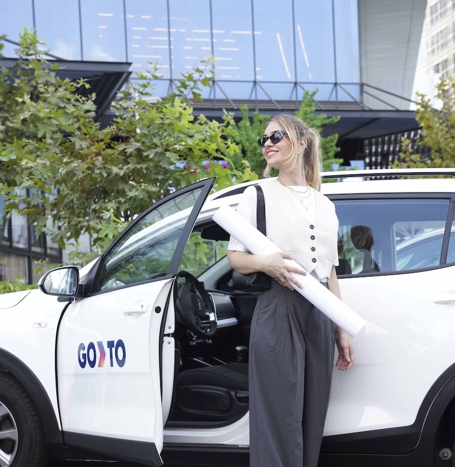 A woman stepping out of a GoTo shared vehicle while holding architectural plans. She is dressed in professional attire, wearing sunglasses, and standing next to the open door of the white car with the GoTo logo visible. The background includes modern office buildings and greenery, suggesting a business environment.