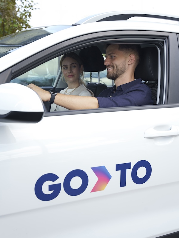 A man and woman sit inside a GoTo-branded car, with the man driving and the woman in the passenger seat. Both are smiling, and the scene captures a casual, pleasant moment. The GoTo logo is prominently displayed on the car door.