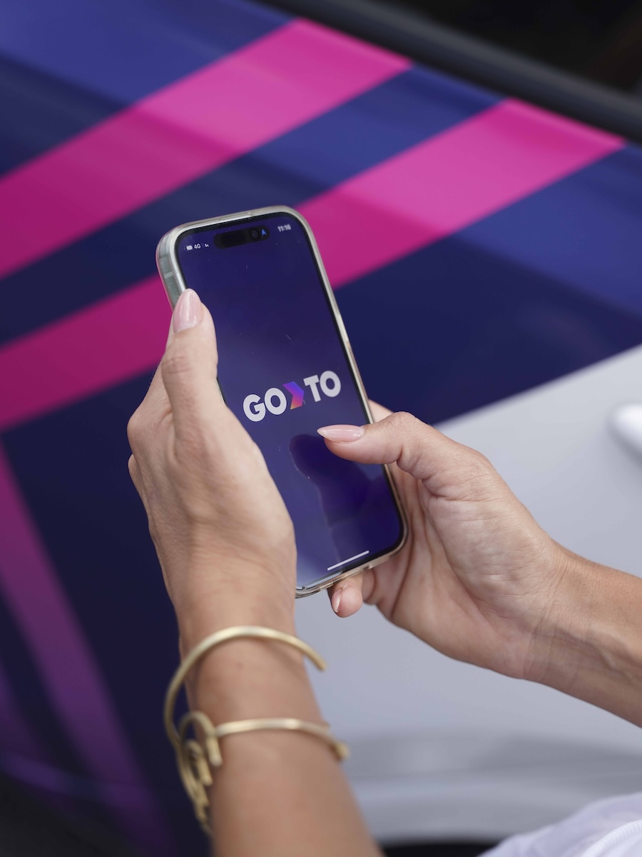 A person holding a smartphone with the GoTo app open on the screen. The background shows a vehicle with diagonal pink and purple stripes, while the phone displays the GoTo logo.