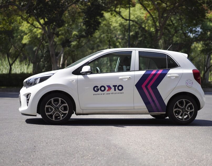 A white compact car with GoTo branding parked in an open area. The vehicle features the "GoTo" logo on the side, along with bold pink and purple arrow graphics.