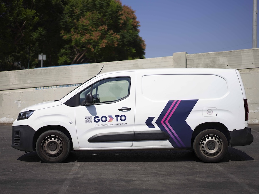 Side view of a white GoTo van rental service vehicle, featuring the company logo, a QR code, and vibrant purple and blue arrows on the side, parked outdoors against a concrete wall.