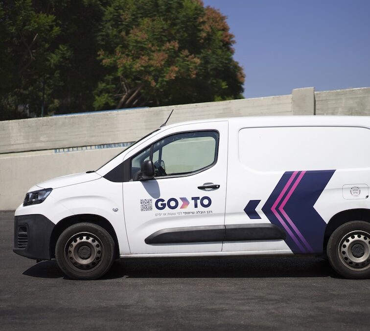 A white GoTo-branded cargo van parked on a paved road, featuring the "GoTo" logo and a bold arrow design in purple and pink. The van is part of the GoTo car-sharing service fleet, suitable for transporting goods or larger items