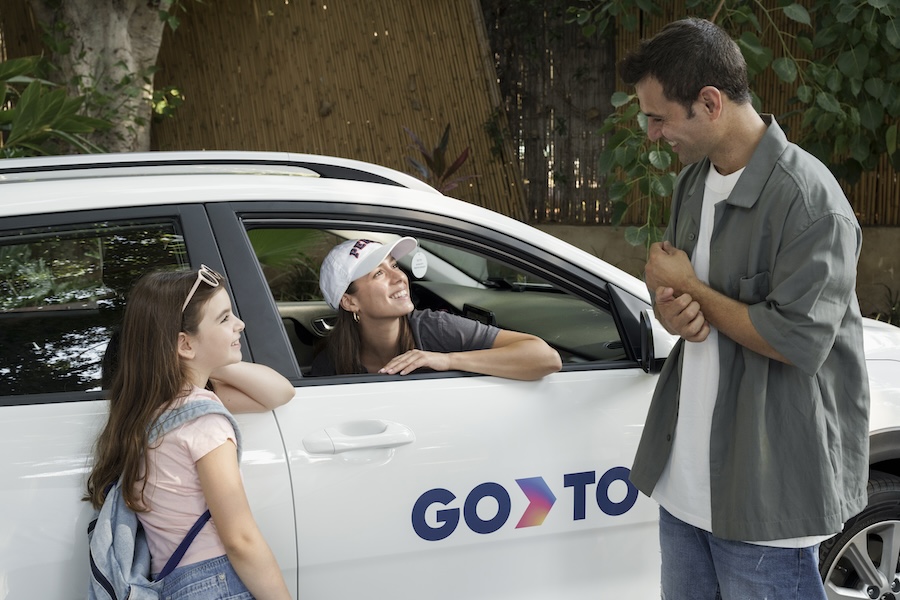 A woman in a GoTo-branded car interacts with a man and a young girl standing outside. The car displays the GoTo logo, and the scene suggests a friendly and casual conversation. The setting includes greenery and a relaxed outdoor environment, highlighting the convenience and family-friendly nature of the GoTo car-sharing service.