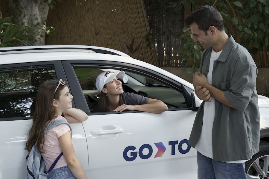 A family interacting near a GoTo car-sharing vehicle. A smiling woman sits inside the car, while a man and a young girl with a backpack stand outside, engaging in conversation. The scene highlights the convenience and accessibility of GoTo's long-term car-sharing service.