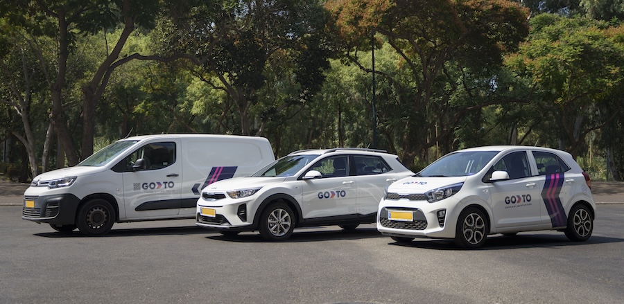 A lineup of GoTo car-sharing vehicles parked in an open area surrounded by trees. The fleet includes a van, a compact SUV, and a small hatchback, all featuring GoTo branding.