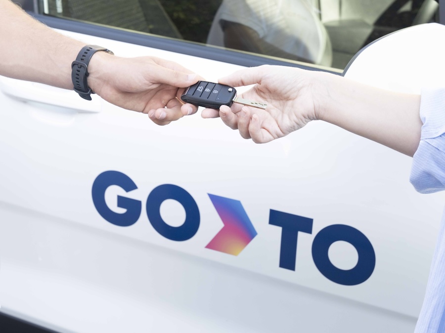 Close-up of two people exchanging car keys in front of a GoTo-branded car, highlighting an efficient car rental for business transaction.